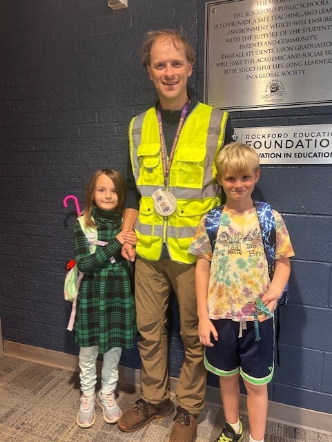 Dad stands wearing a bright yellow reflective vest with two students.